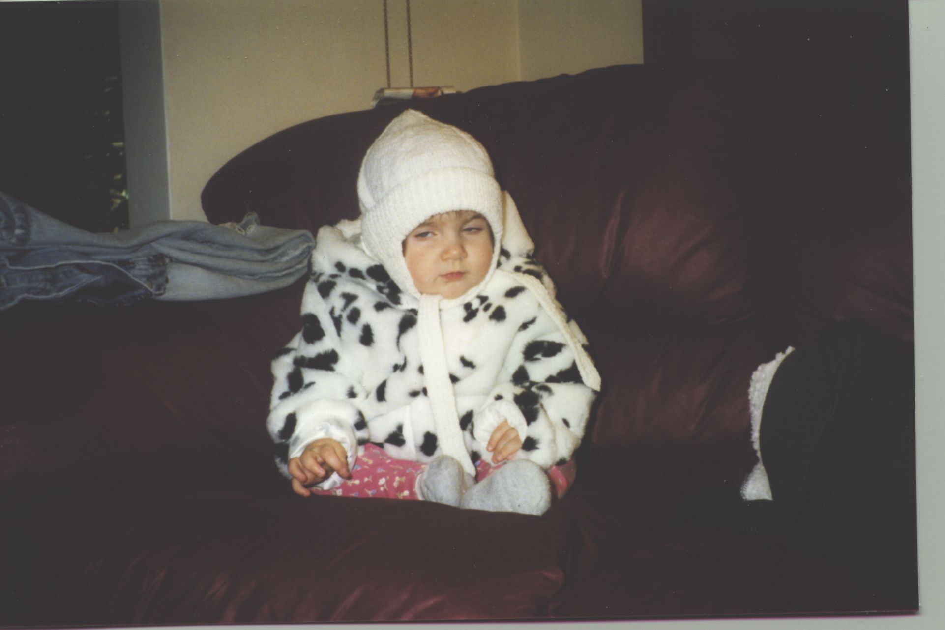 A one-year-old Night sits on a dark burgundy sofa. He is wearing a white hand-knit earflap hat, fluffy baby blue booties, magenta leggings with a small yellow floral print, and a black and white cow-print jacket. He has an unidentifiable expression on his face, eyes narrowed as if mid-blink.